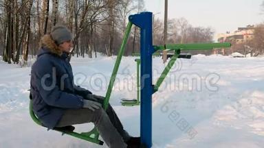 穿着带罩的蓝<strong>羽绒服</strong>的人正在<strong>冬季</strong>公园的模拟器上做腿部运动。 侧视。